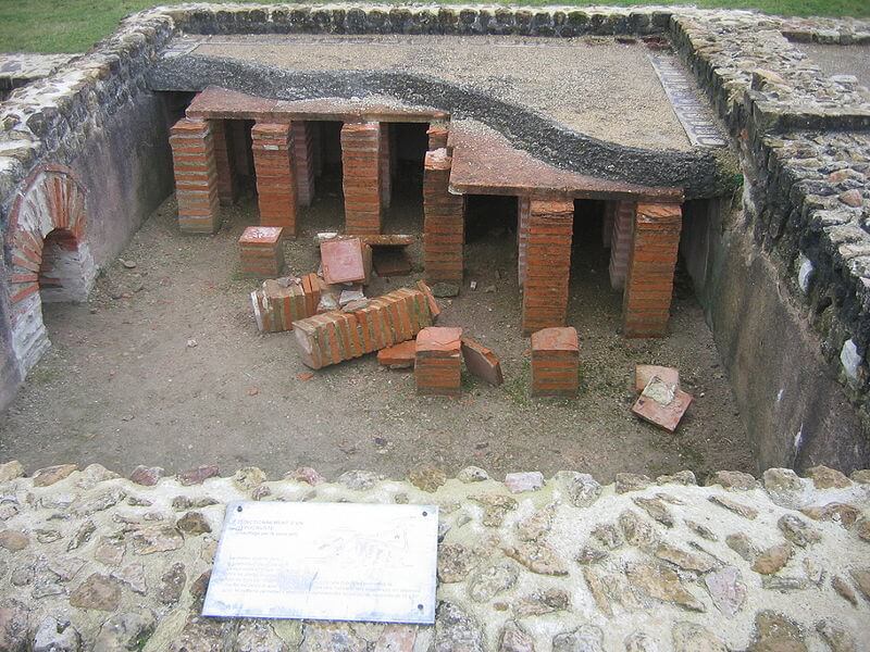 Photograph of a hypocaust with part of the floor missing. The remaining area of floor is held up by stacked brick tiles. On the left a hole in the wall would have let in hot air from a furnace.